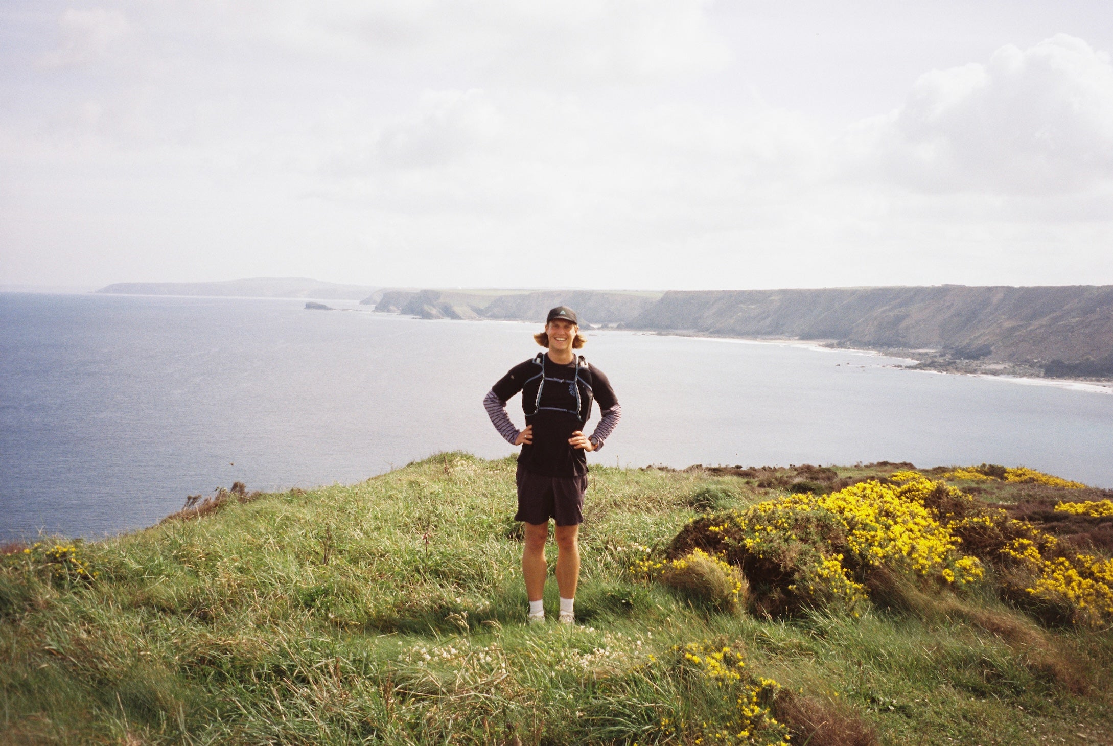 Person standing on a grassy hill overlooking a body of water with mountains in the distance.