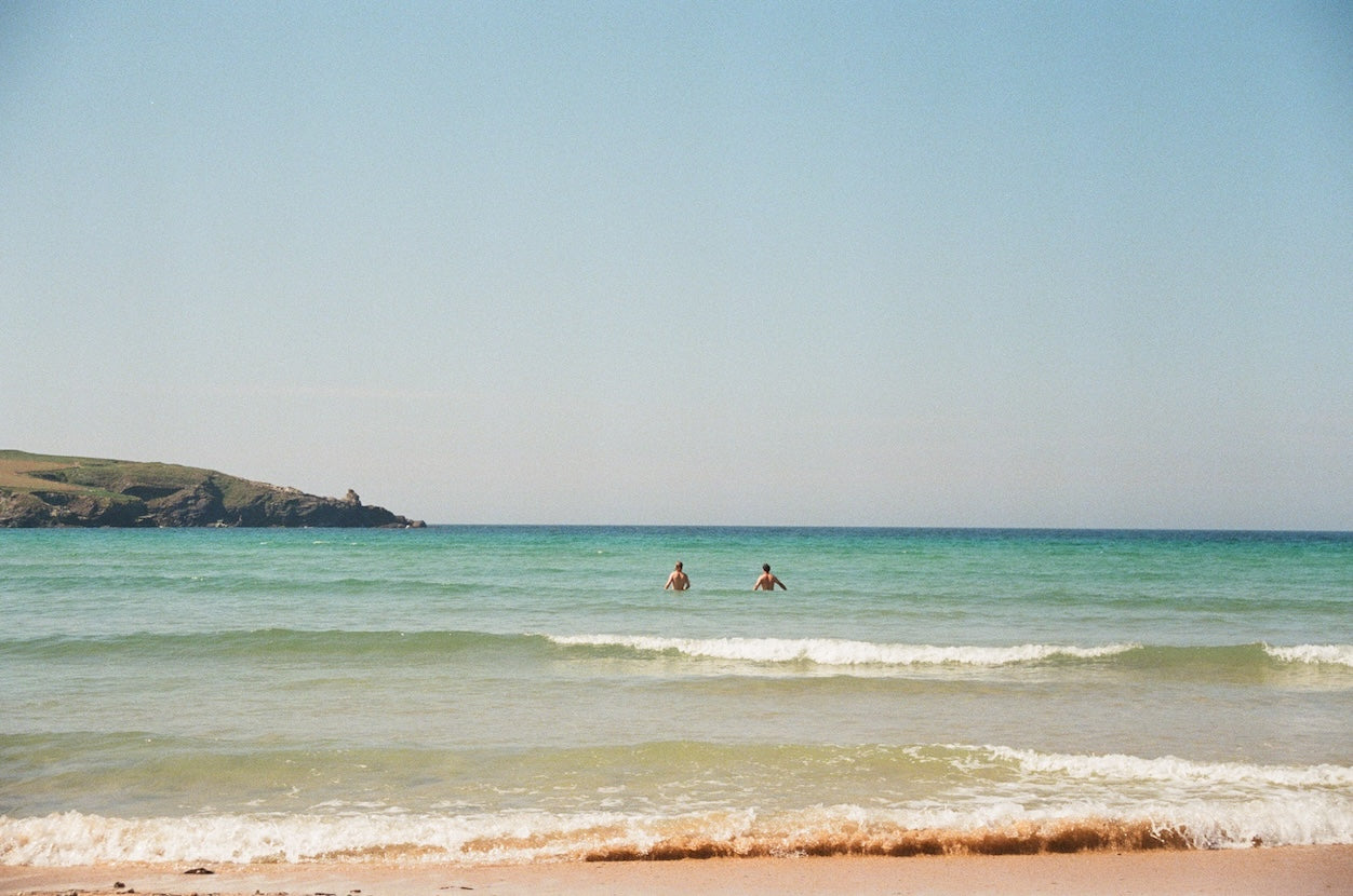 Two people swimming in a calm ocean with a clear blue sky and cliffs in the background.