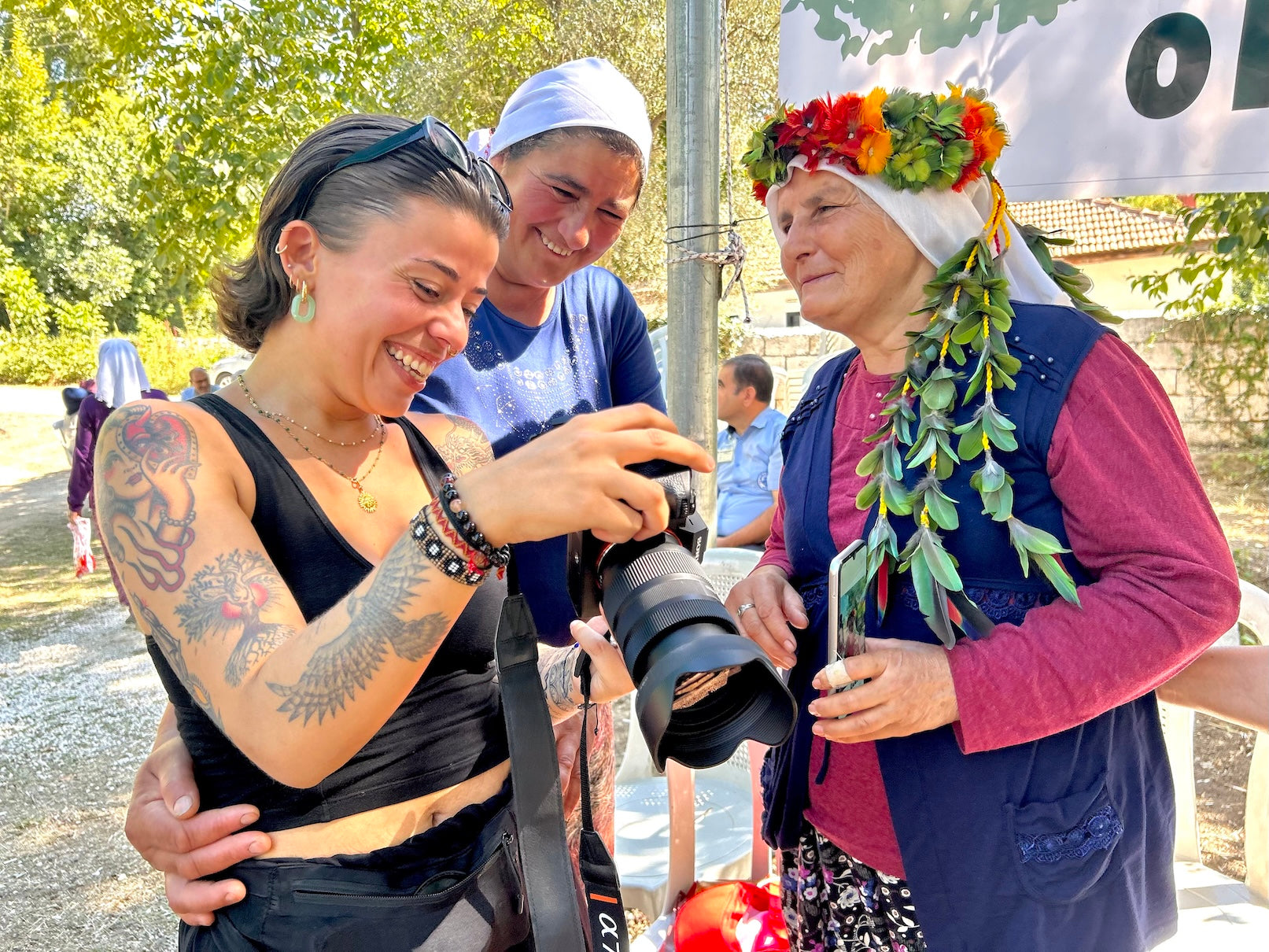 Two people taking a photo of an elderly woman with a floral headpiece outdoors.