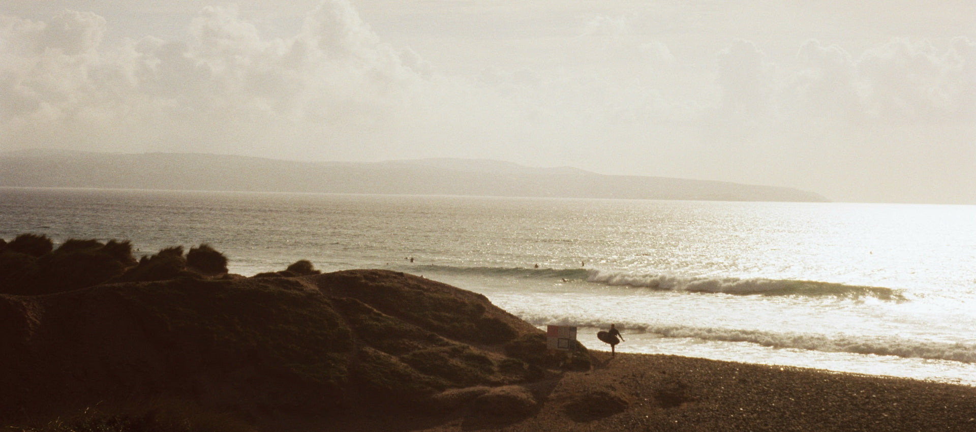 A film photo of the coast in Cornwall with a surfer carrying a board walking along the beach