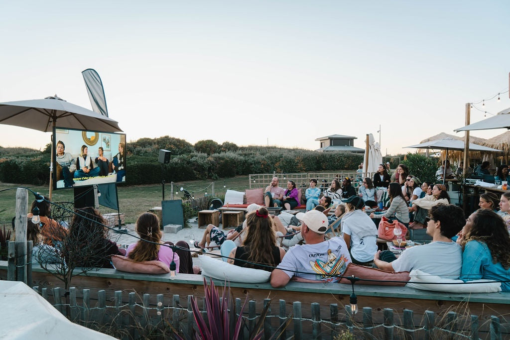 A large group of people sat around outside watching a surf film at dusk on a big outdoor screen