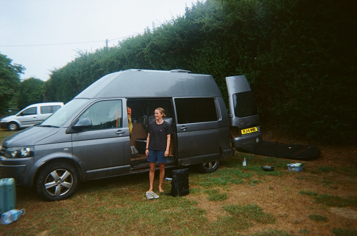A gray camper van parked on grass with its side door and rear hatch open, with a person standing outside the vehicle. Trees and a second vehicle are visible in the background.